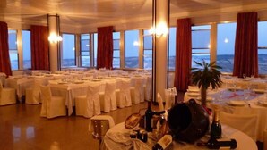 A banquet hall with round tables set for dining, large windows with curtains, and a view of the sea. - Hotel Astoria (Fermo)
