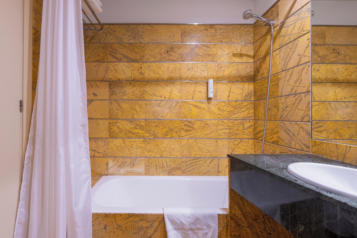 Une salle de bain avec des carreaux de marbre jaune, une baignoire blanche, un plan de toilette noir et une pomme de douche.