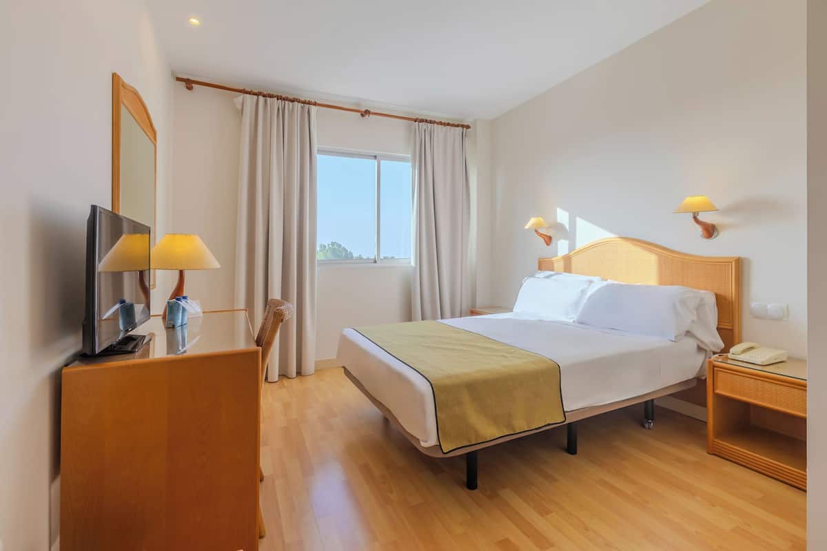 A hotel room with a bed, bedside lamps, a TV, a wooden desk, and a window with curtains.