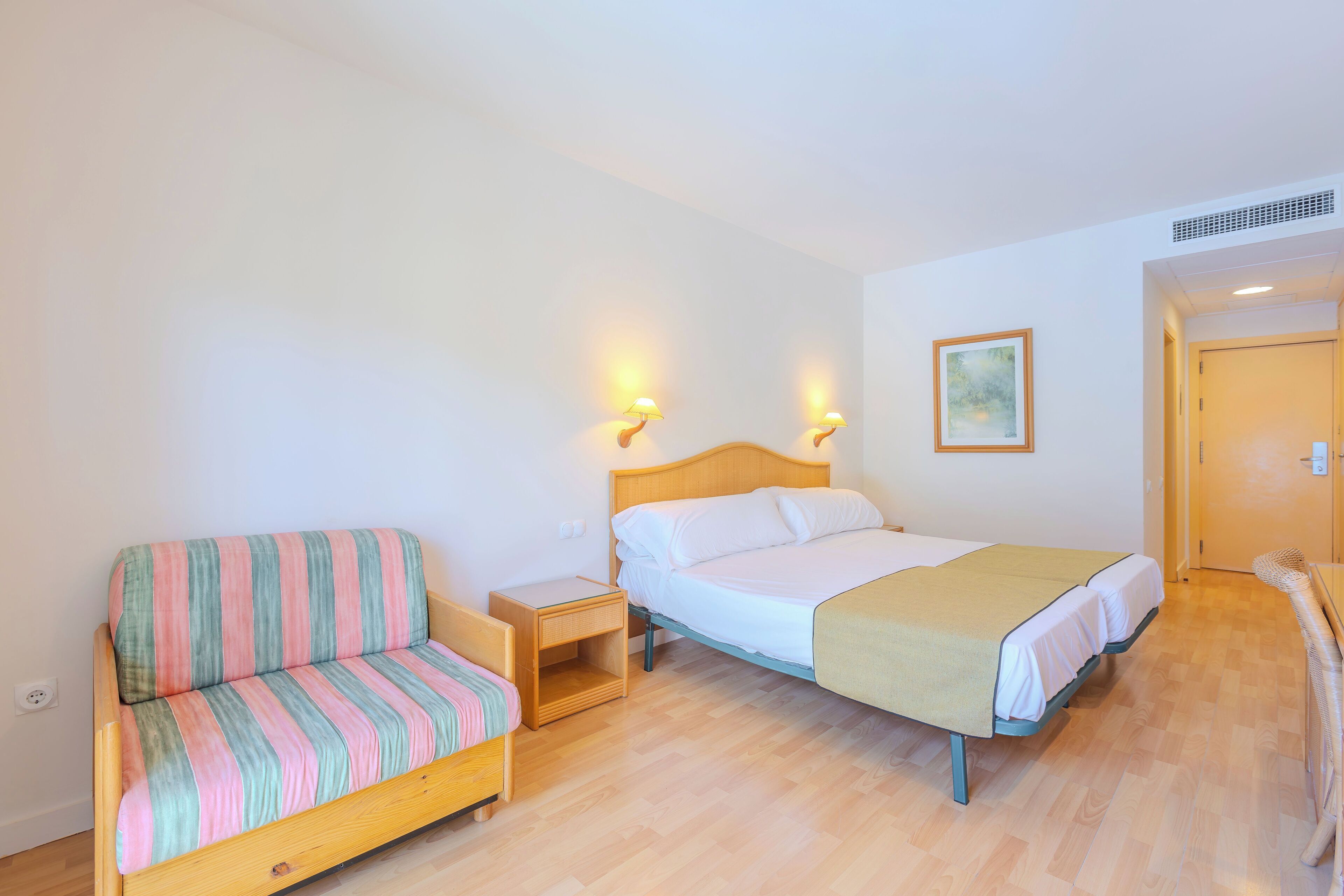 Un dormitorio con cama, sofá a rayas, mesita de noche y lámpara de pared.
