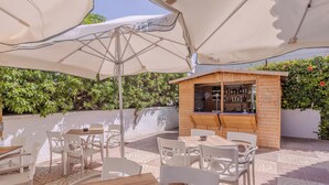 Une terrasse extérieure avec des chaises et des tables blanches, un coin bar en bois et de grands parasols pour créer de l’ombre.