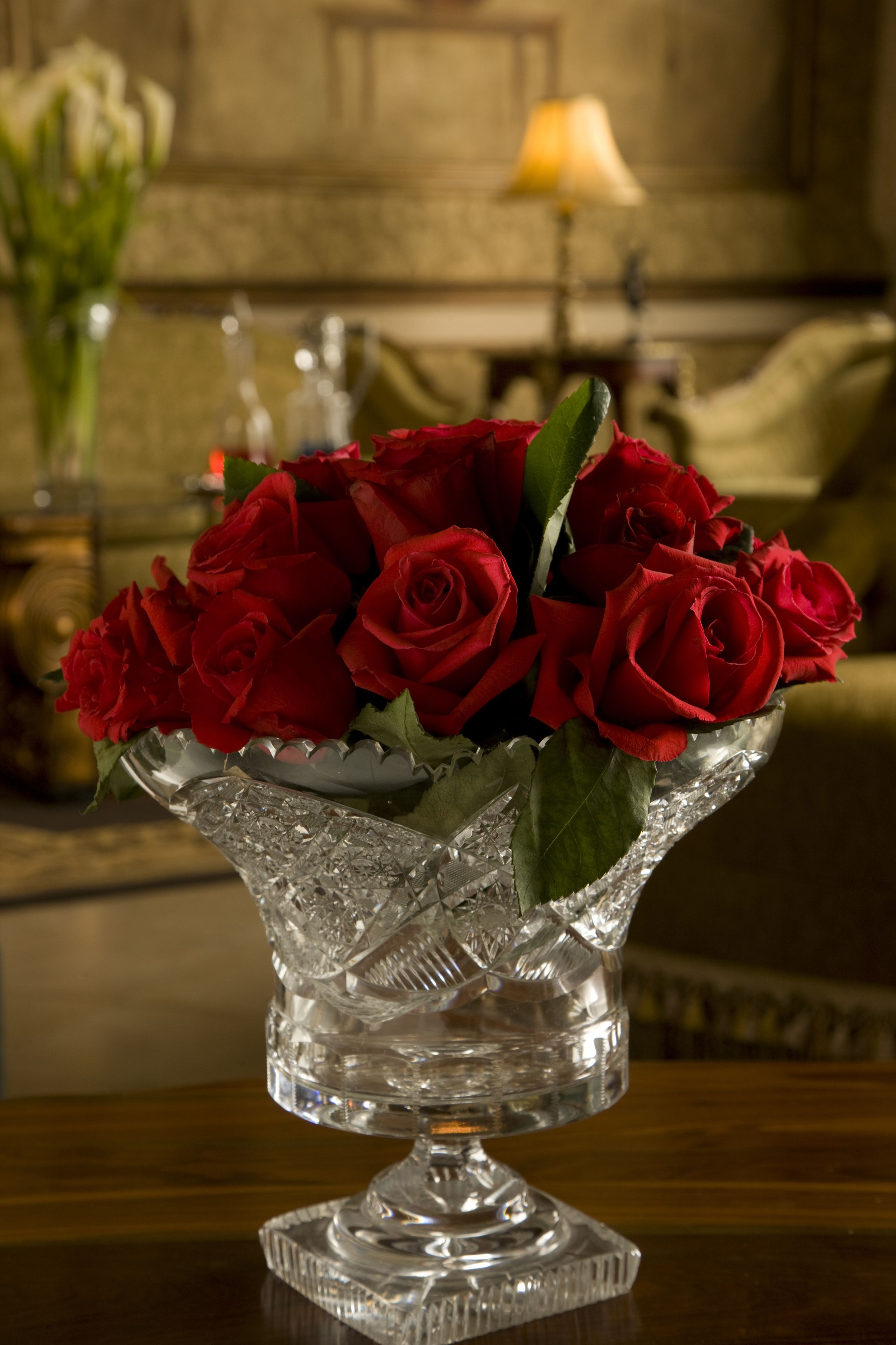 A clear glass vase with red roses on a wooden table in a room with a lamp and a couch in the background.