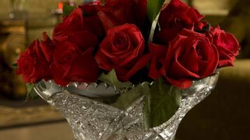 A clear glass vase with red roses on a wooden table in a room with a lamp and a couch in the background.