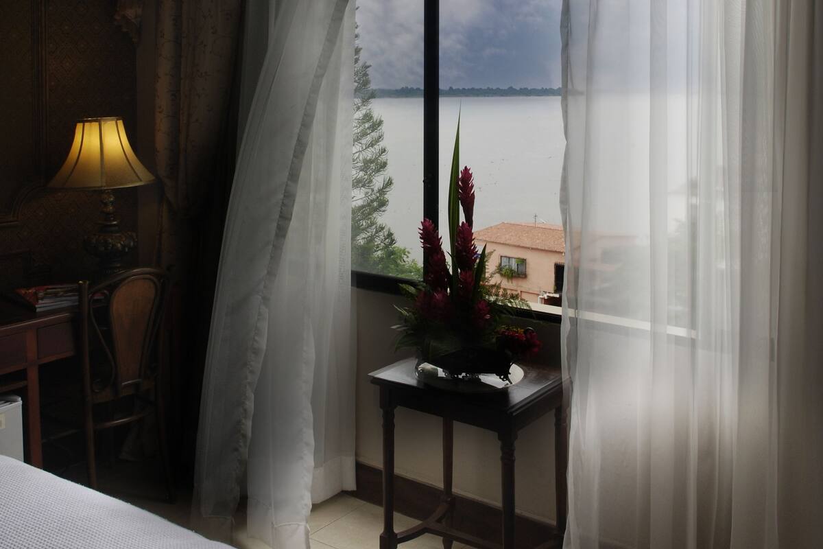 A hotel room with a large window, a bedside table with a lamp, and a view of the sea.