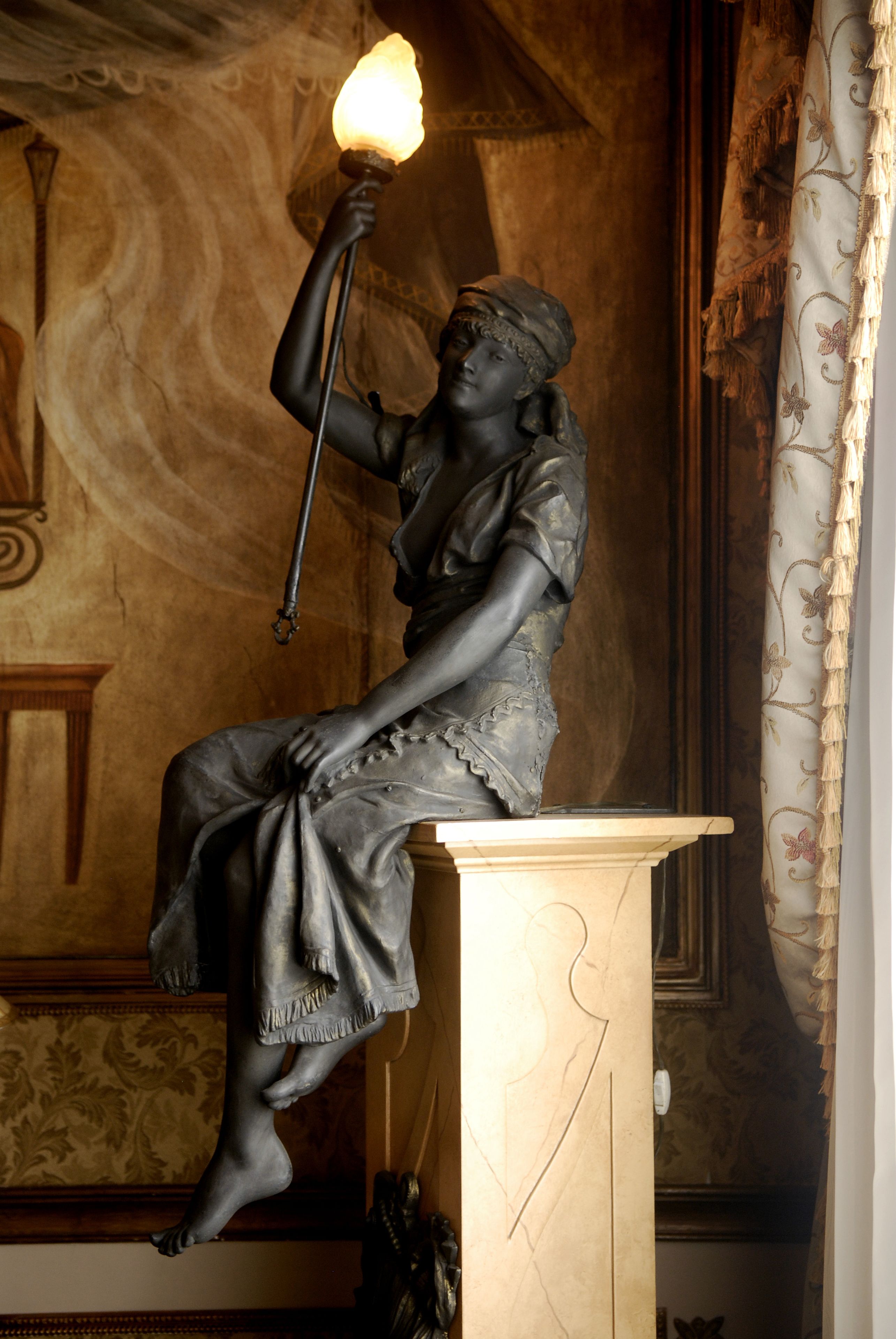 Una estatua de una figura sentada sosteniendo una antorcha, ubicada sobre un pedestal en una habitación con papel tapiz estampado y un aparador de madera.