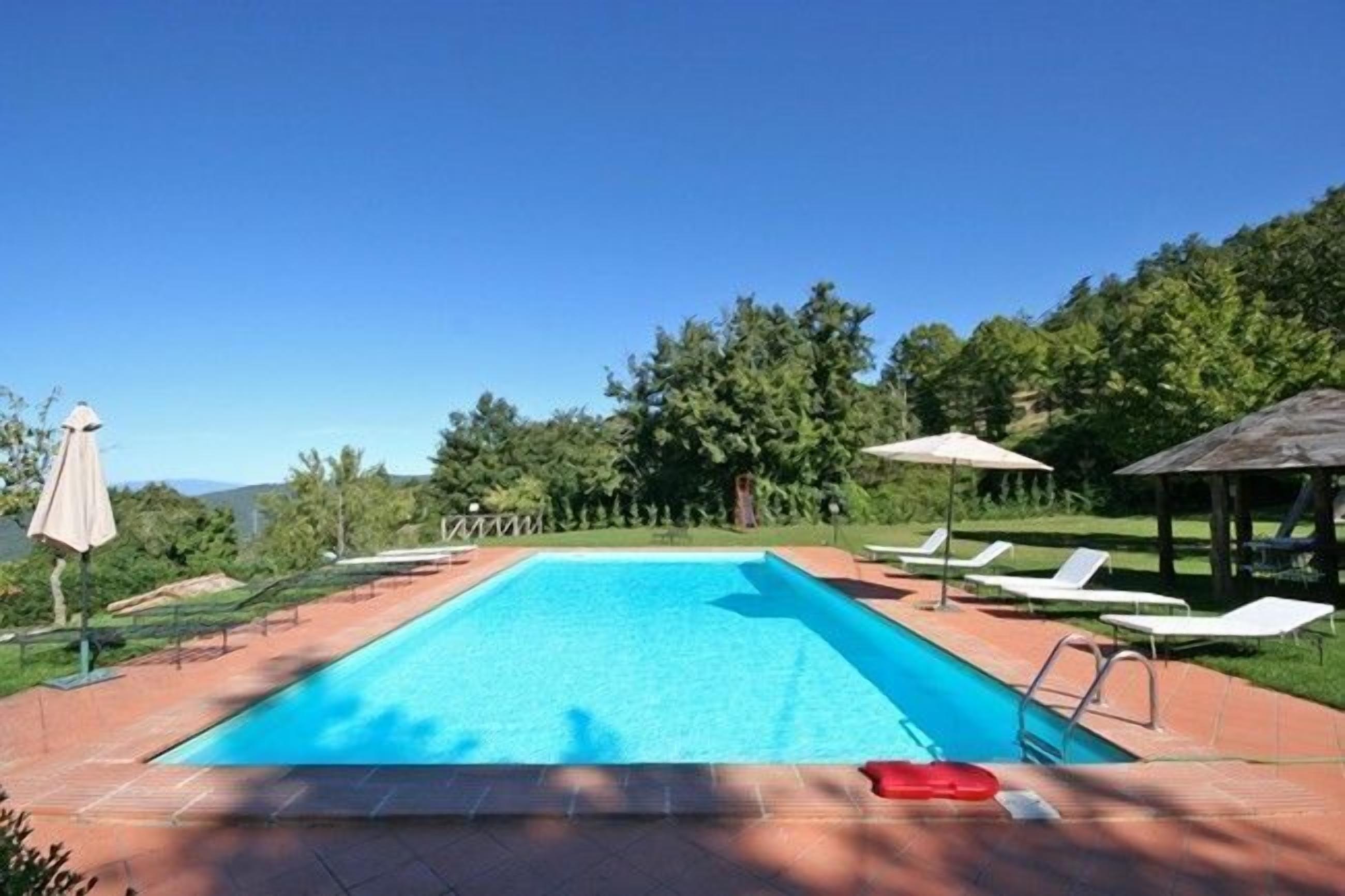 Una piscina rectangular con superficie de agua azul cristalina, rodeada por una terraza con baldosas. Se observan sillas de descanso y sombrillas alrededor de la piscina.