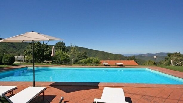 A swimming pool with lounge chairs and an umbrella, surrounded by greenery and hills. - Villa Margherita Due (Cortona)