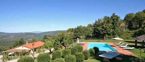 Ein Poolbereich mit Liegestühlen und Sonnenschirmen, umgeben von ordentlich geschnittenen Büschen und Bäumen, mit Blick auf die Berge.