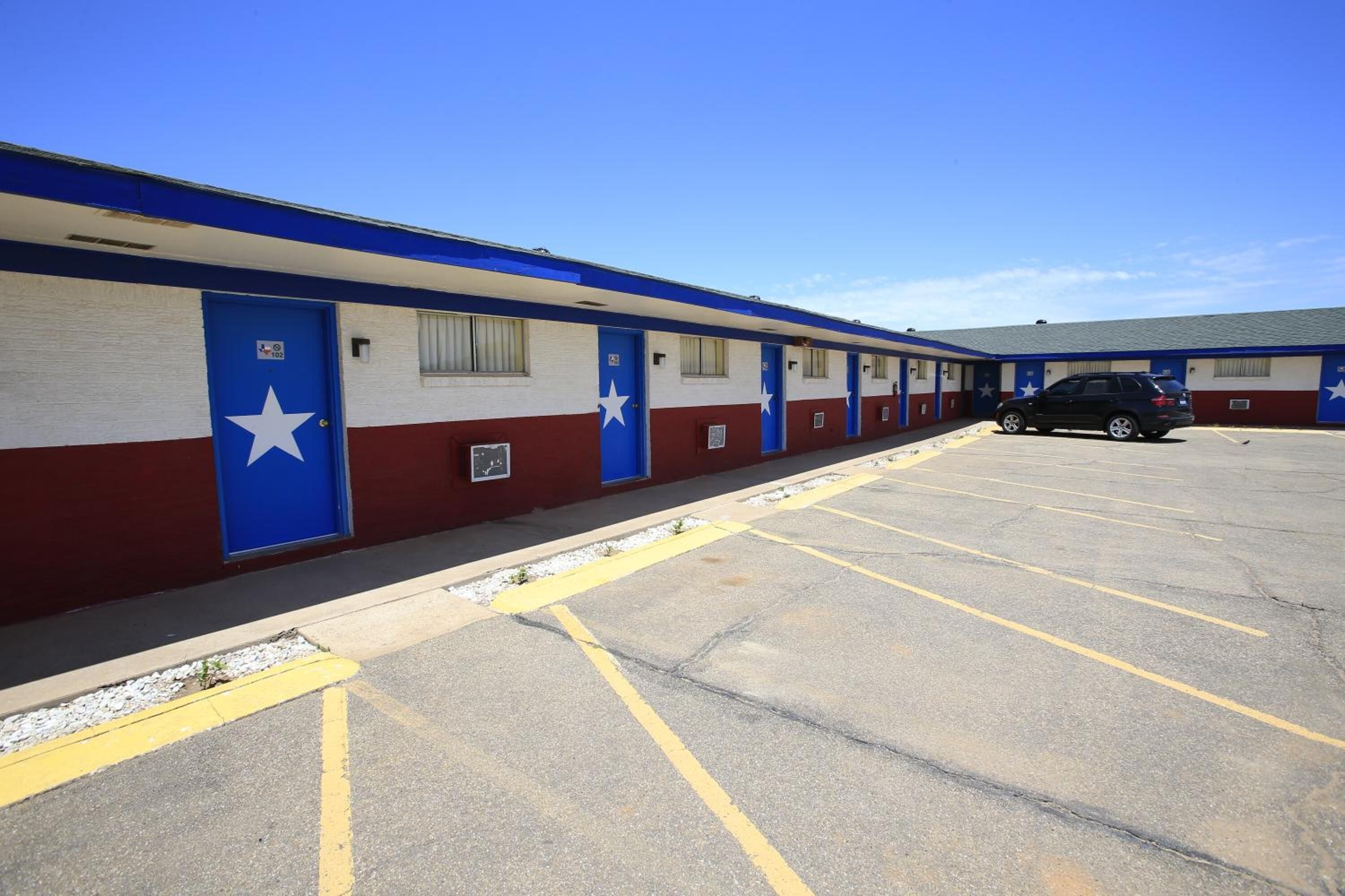 A motel with a row of rooms featuring blue doors with white stars, a parking lot, and a clear sky.