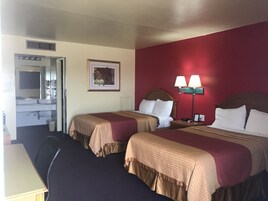 Habitación de hotel con dos camas, un escritorio, una silla, un baño con espejo y un cuadro colgado en la pared.