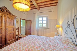 A bedroom with a large bed, wooden wardrobe, a chair, a window with curtains, and a lamp on a bedside table. - Villa Leonardo (Vinci)