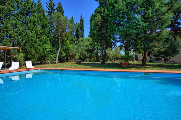 A clear blue swimming pool with white lounge chairs, surrounded by trees and a well-maintained lawn.