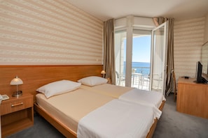 A hotel room with a large bed, a desk with a telephone, and a view of the sea through an open door.