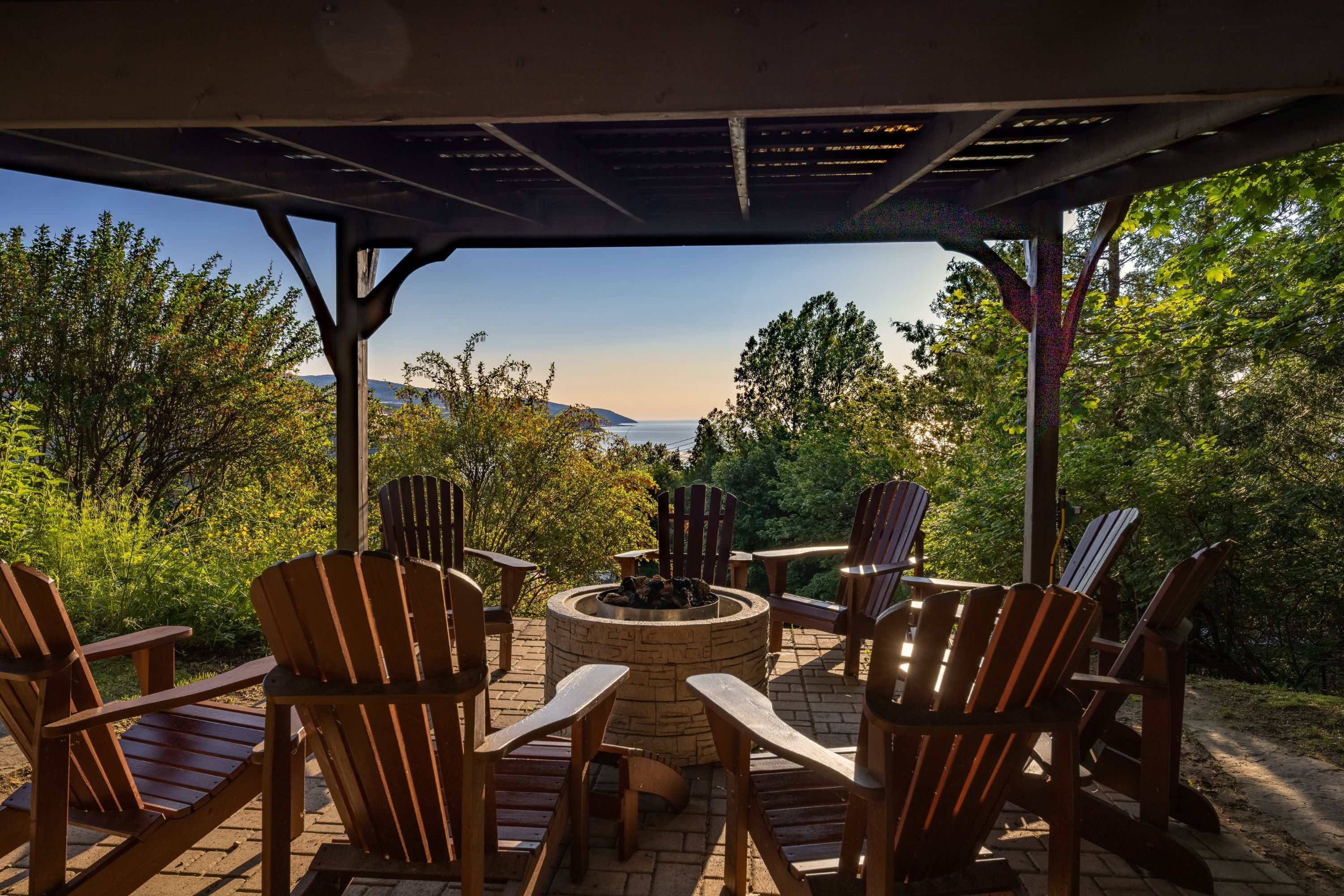 An outdoor seating area with Adirondack chairs and a fire pit, surrounded by trees and a view of the landscape.