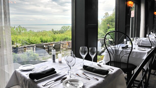 Une table à manger dressée avec une nappe blanche, des assiettes, des couverts et des verres à vin, offrant une vue panoramique sur un paysage verdoyant et une étendue d’eau.
