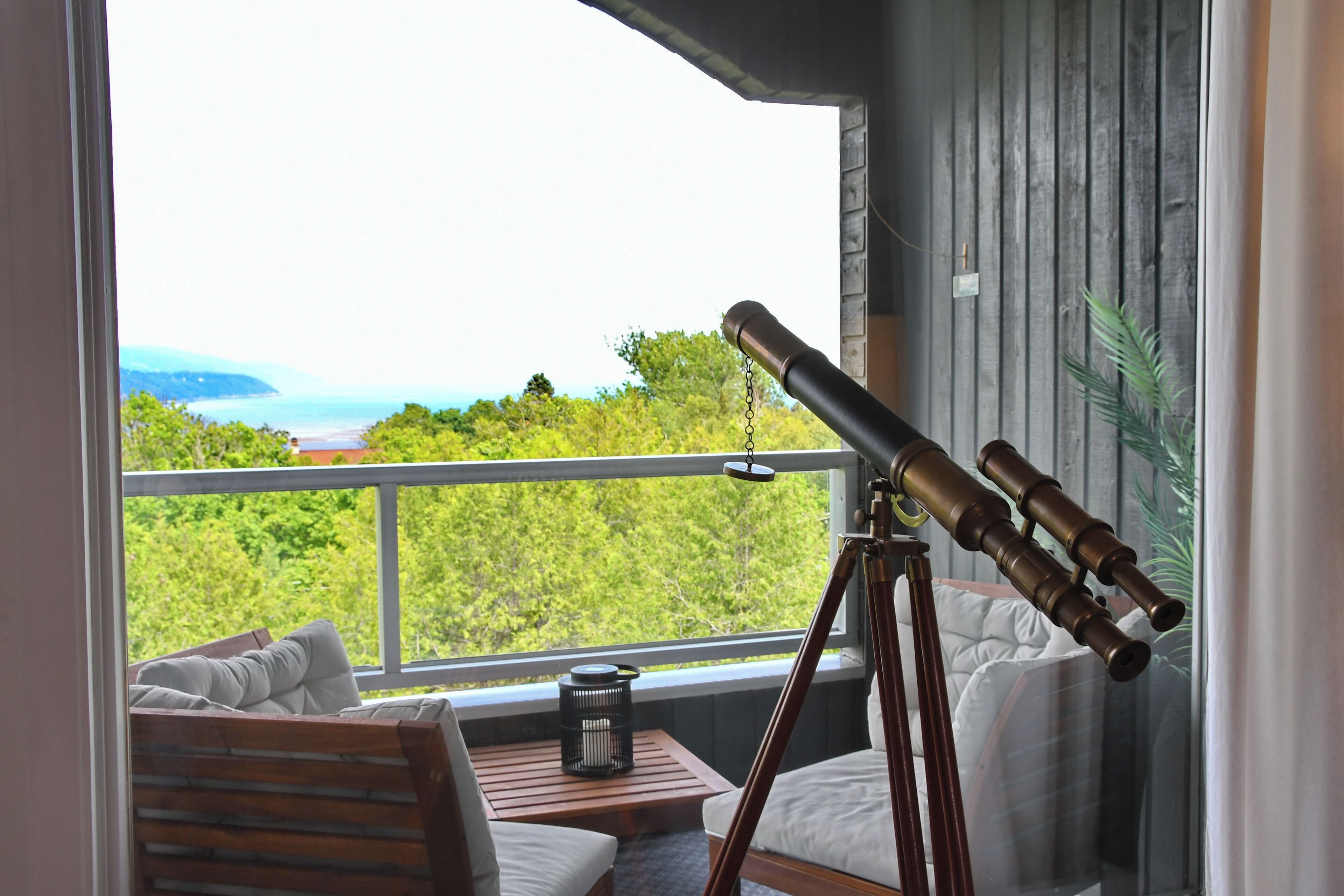 A balcony with a telescope, a wooden chair, a small table, and a view of the sea and greenery.