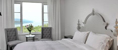 A hotel room with a large bed, two armchairs, a small table with a vase, and a view of the sea through a window.