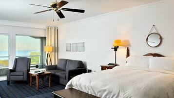 A hotel room with a bed, two armchairs, a desk, a ceiling fan, and a view of the ocean.