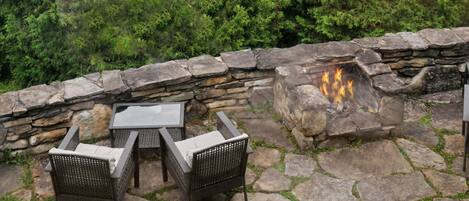 An outdoor seating area with stone flooring and a fire pit, surrounded by lush greenery and overlooking a forested landscape.