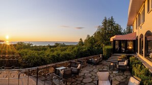 Une terrasse en pierre avec des meubles d’extérieur, une vue sur la mer et un bâtiment doté de grandes fenêtres.