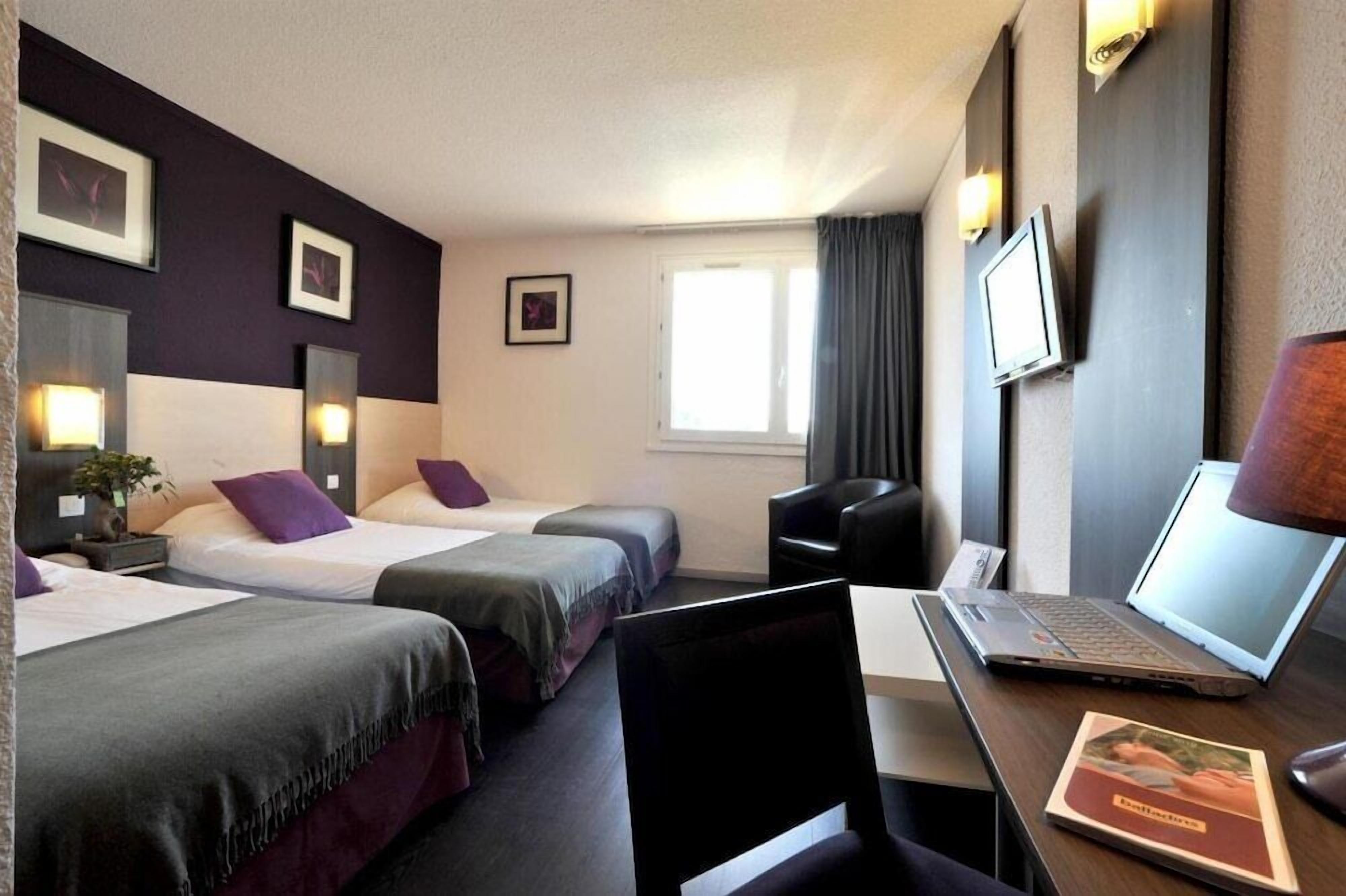 A hotel room with two beds, a desk with a laptop, a chair, a TV, and framed pictures on the wall.