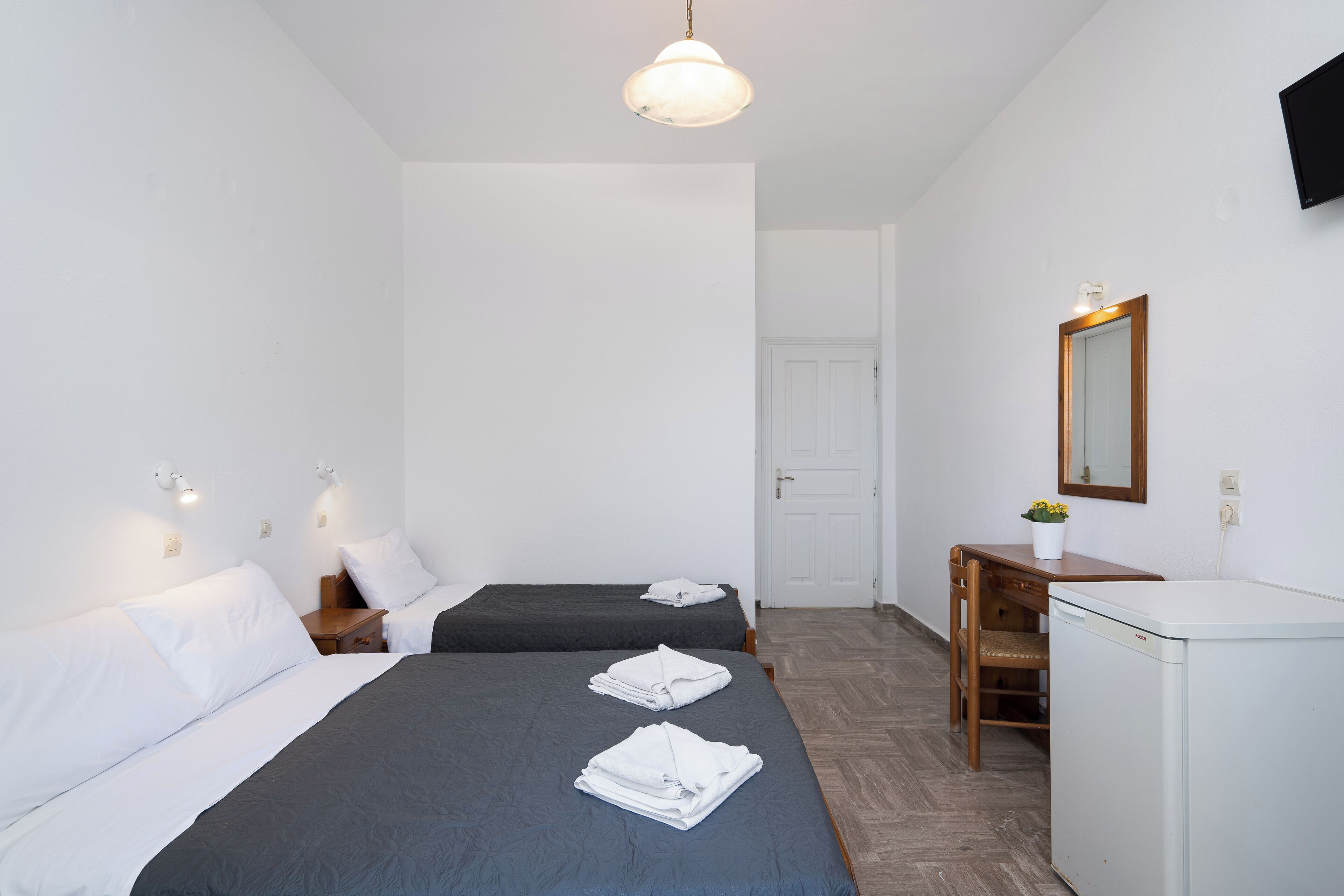 Habitación de hotel con dos camas individuales, un escritorio, una silla, un refrigerador pequeño y un televisor montado en la pared.