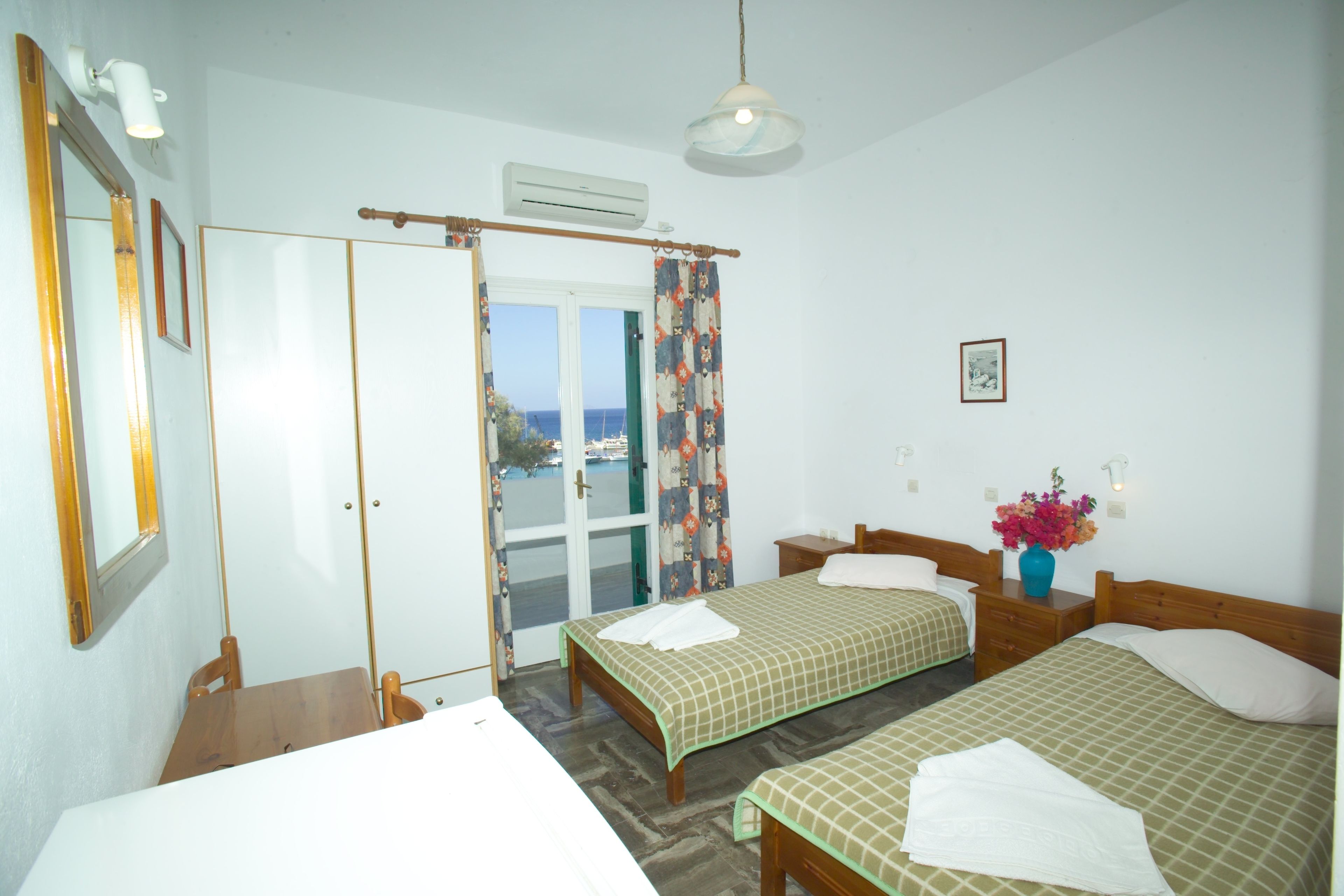 A hotel room with two beds, a wooden nightstand, a small table, a mirror, and a view of the sea through the door.