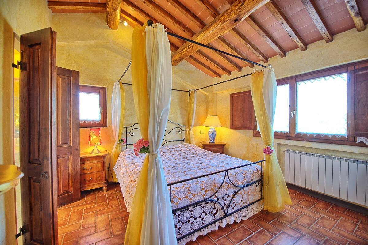 Ein Schlafzimmer mit Himmelbett, Holzbalken an der Decke, einem Fenster mit Blick und einem Nachttisch mit Lampe sowie einem Heizkörper.