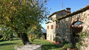 A stone building with a rustic exterior, a garden with a table and chairs, and a tree providing shade. - Casa Anna (Barberino Tavarnelle)