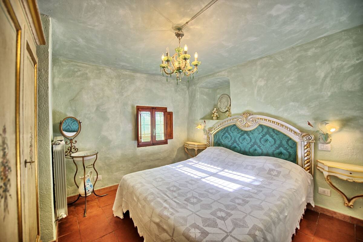 Ein Schlafzimmer mit einem Bett, einer Chandelier, einem kleinen Tisch, einem Spiegel und einem Fenster.