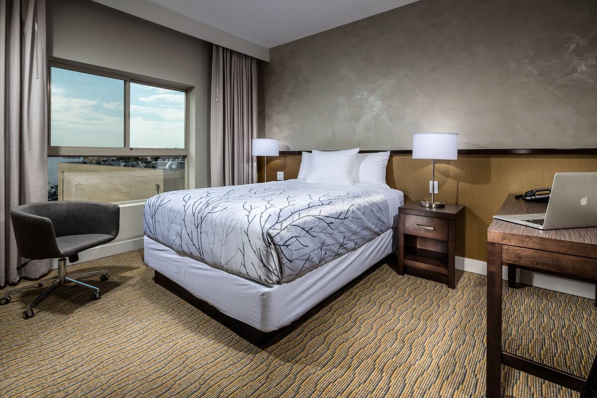 A hotel room with a large bed, a desk with a laptop, a chair, and a window with curtains.