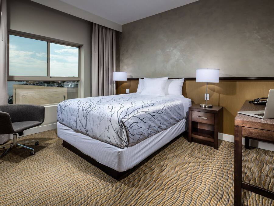 A hotel room with a large bed, a desk with a laptop, a chair, and a window with curtains.