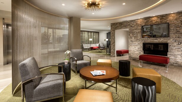A modern living room with a sofa, armchair, coffee table, and ottoman. There is a fireplace and a television mounted on the wall. - Aventura Hotel (Los Angeles)