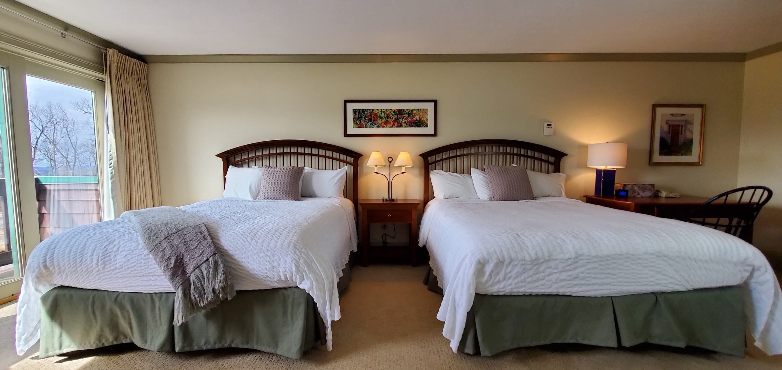 A hotel room with two beds, each with a green skirted base, a headboard, and a white quilted bedspread. There is a window with curtains, a framed picture on the wall, and a nightstand with a lamp.