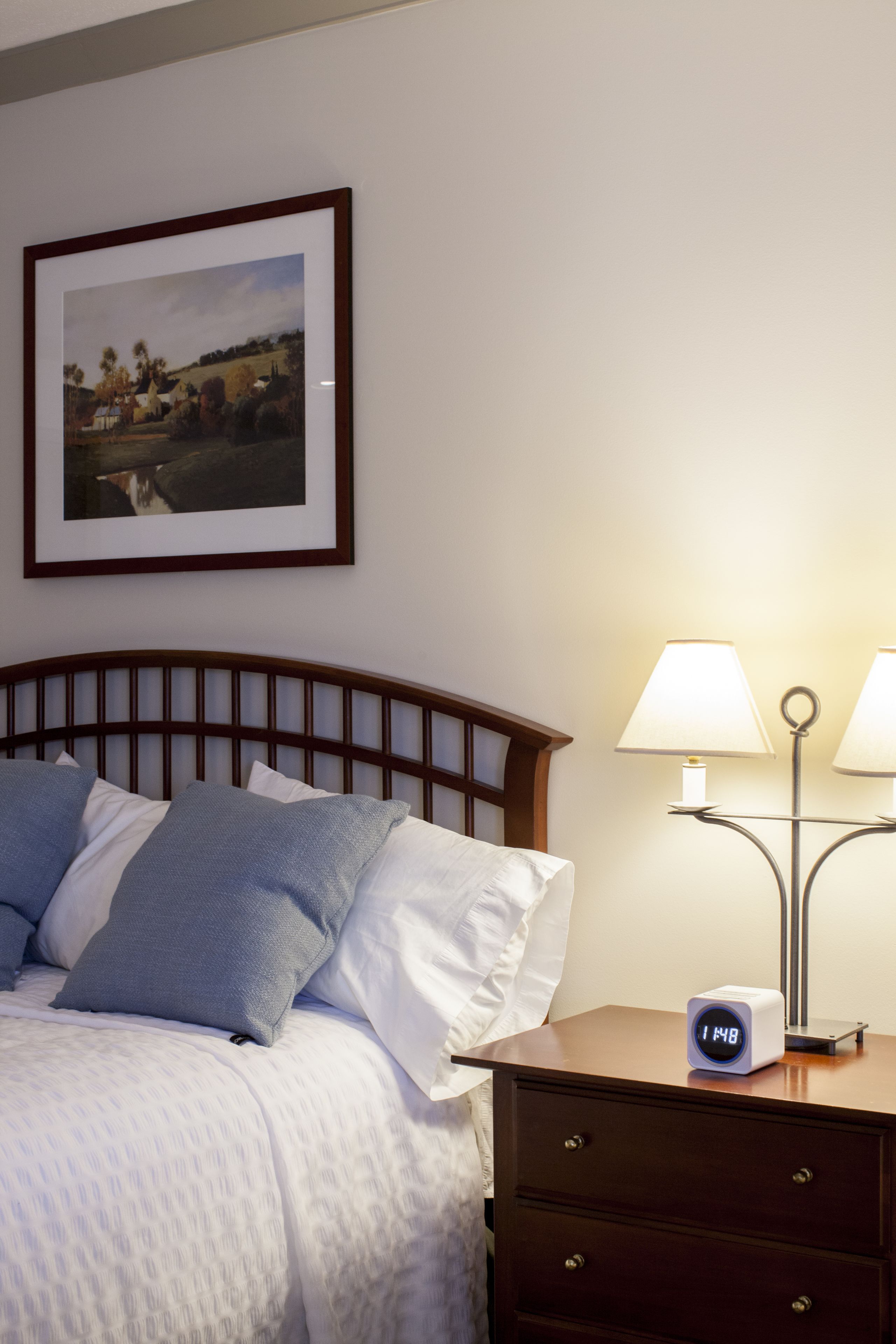 A neatly made bed with pillows, a bedside table with a lamp, a clock, and a framed picture on the wall.