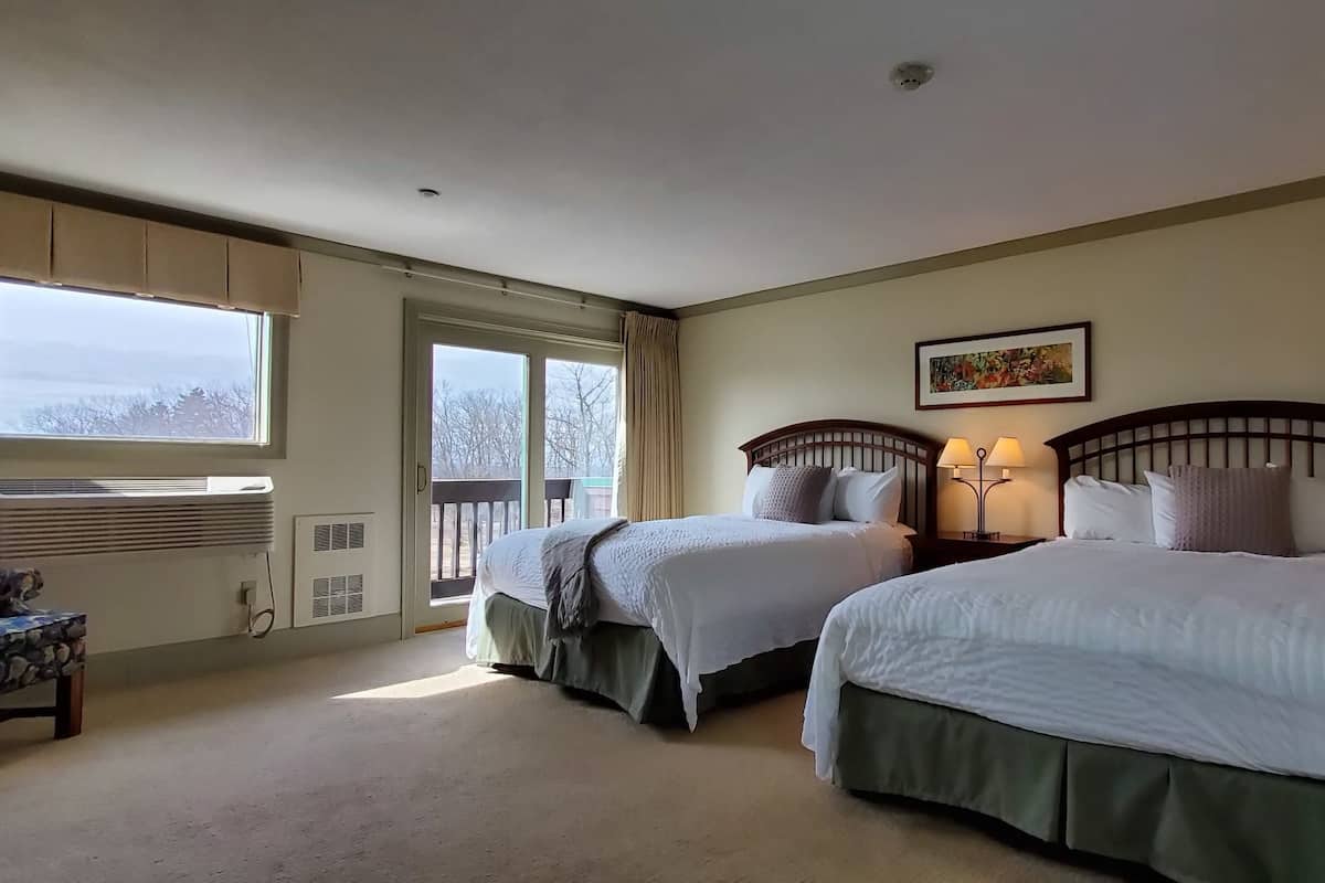 A hotel room with two beds, a window with curtains, a chair, and a bedside table with a lamp.