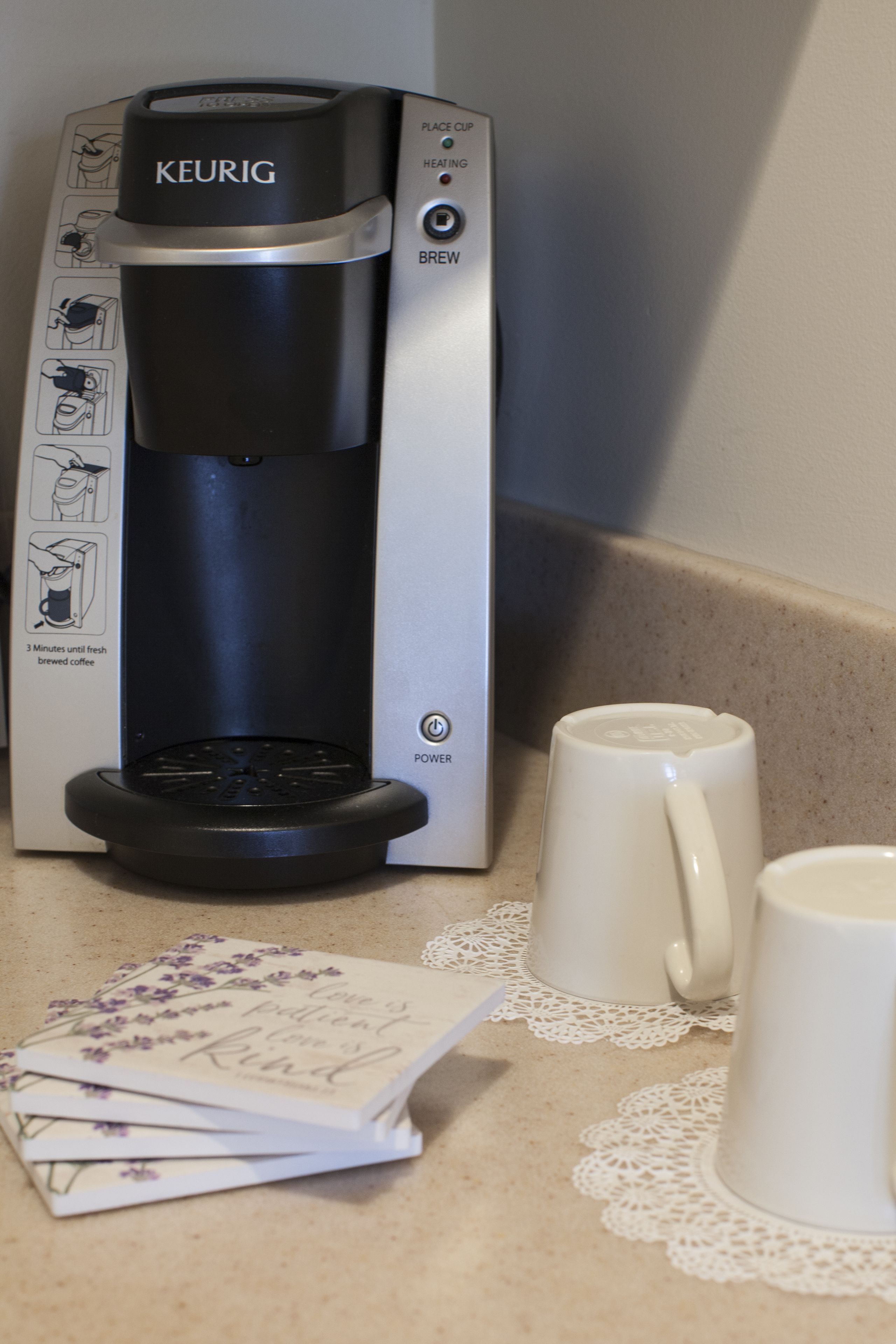 Eine Keurig-Kaffeemaschine auf einer Küchenarbeitsplatte, zusammen mit zwei weißen Tassen und einem Kartenspiel mit floralem Muster.