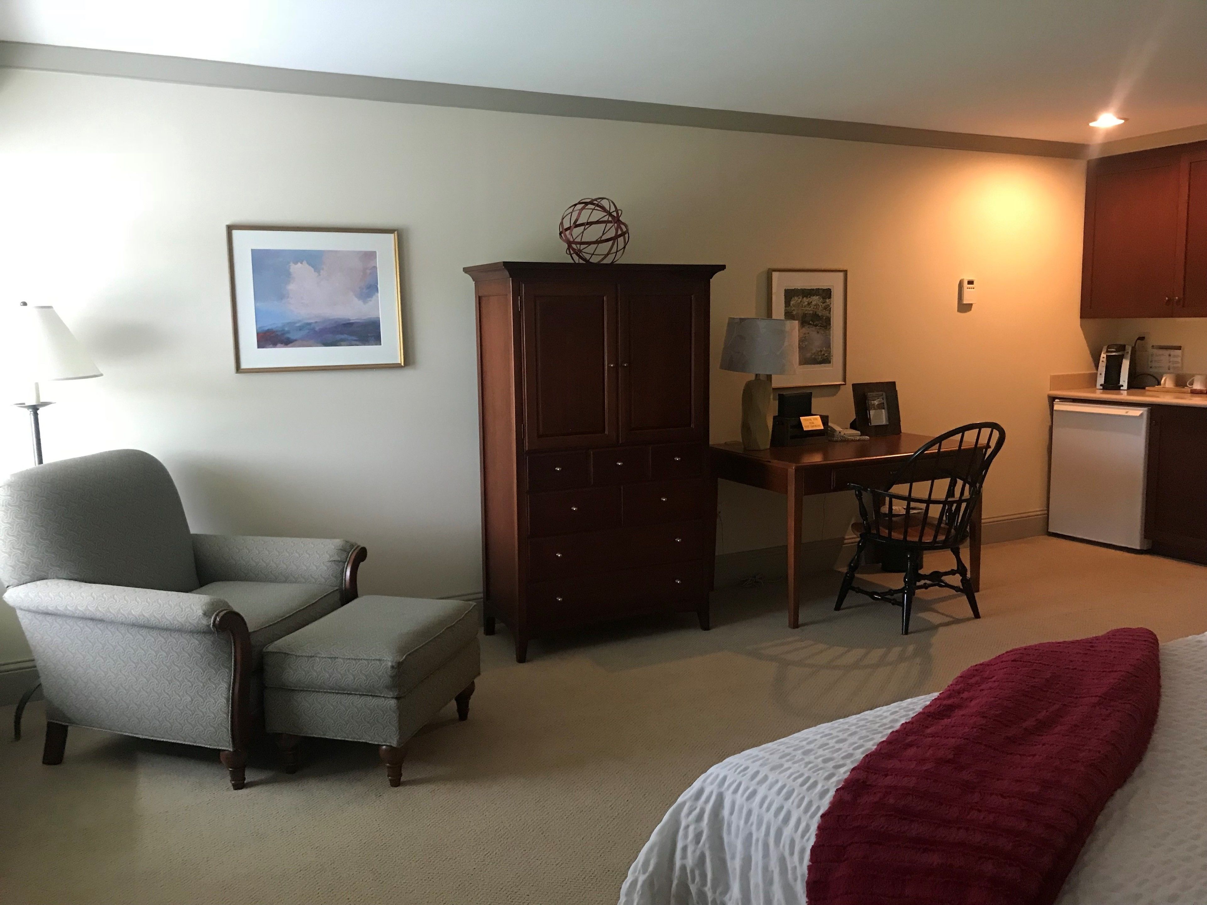 A compact hotel room with a bed, a desk with a chair, a kitchenette, and a large wardrobe.