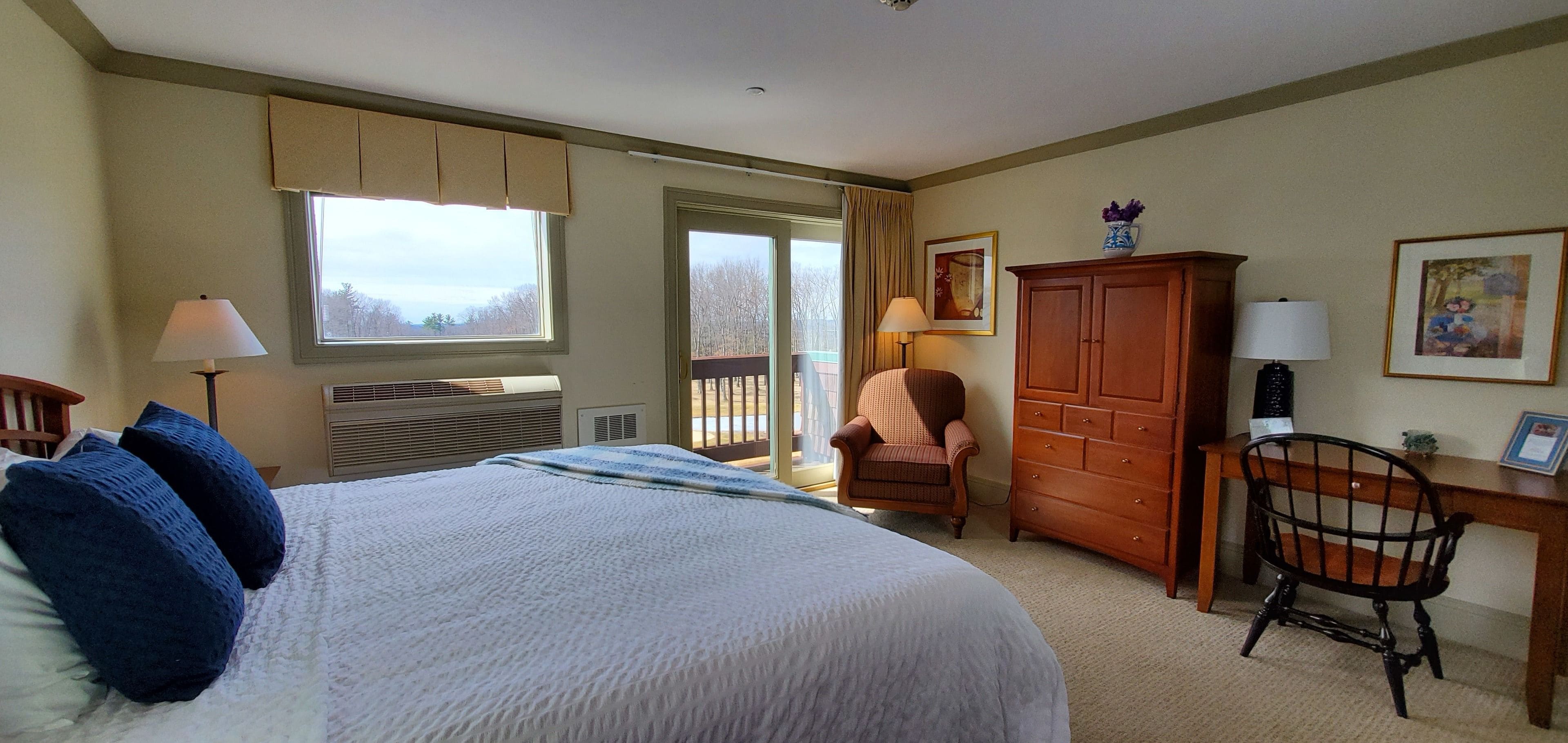A hotel room with a large bed, a desk, a chair, a wardrobe, and a window with a view of trees.