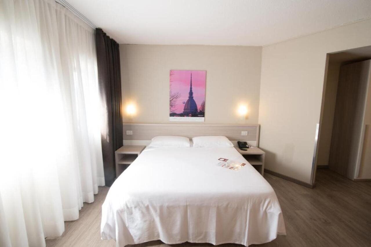Habitación de hotel con una cama grande, dos mesitas de noche, una lámpara de pared y un cuadro de un edificio.