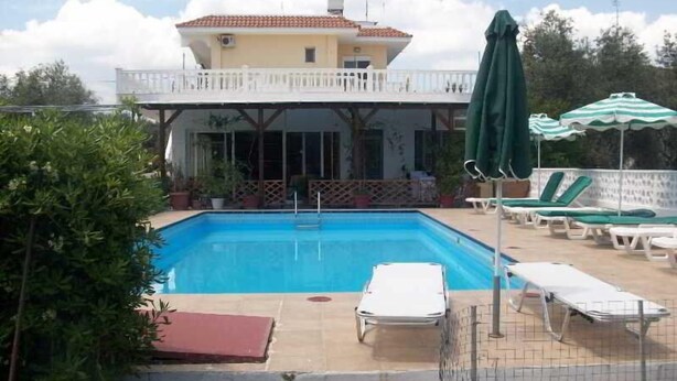A swimming pool with lounge chairs and umbrellas, surrounded by a fence and greenery. - Varelis (Rhodes)