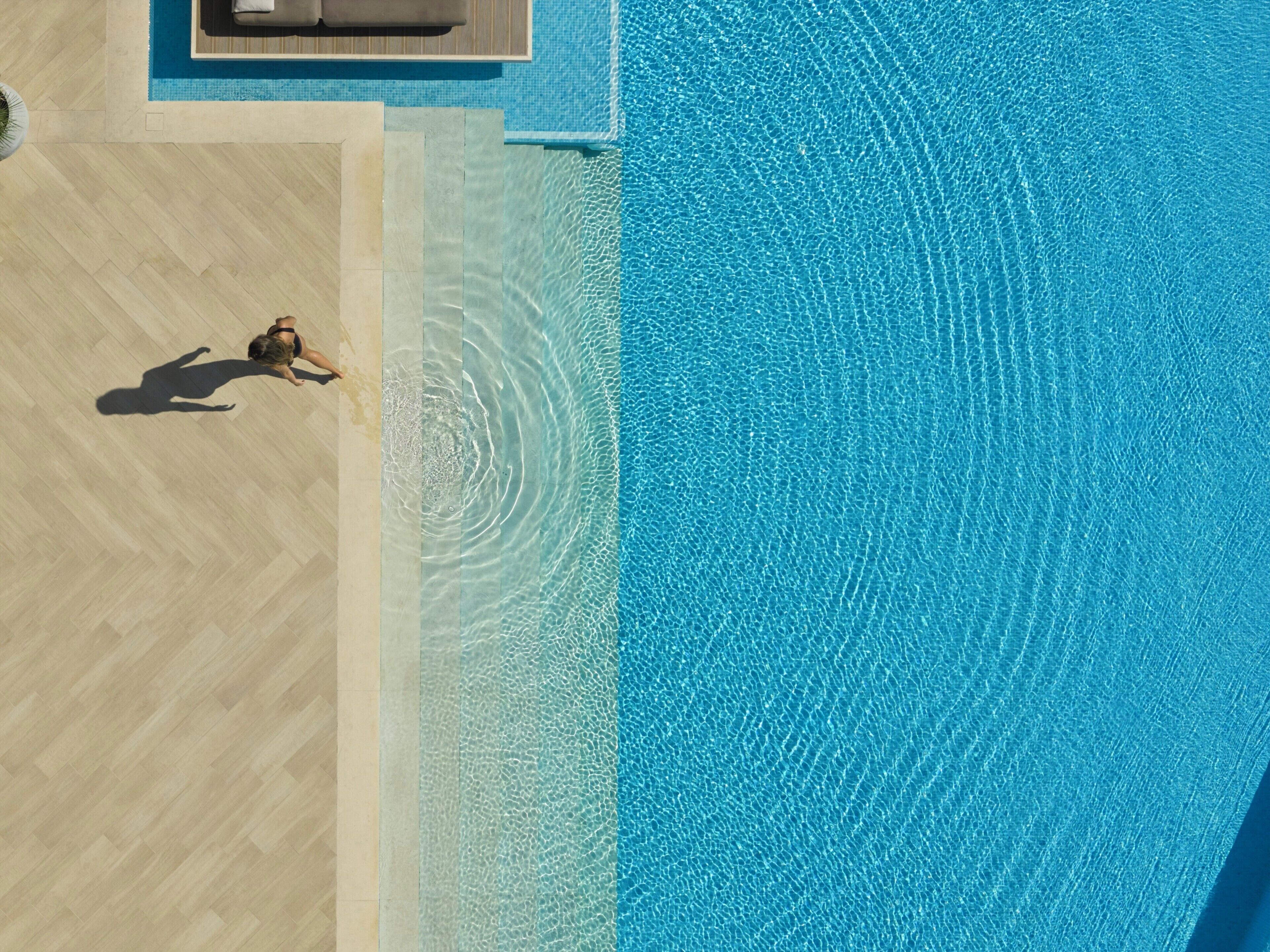 Vista aérea de una piscina con una persona recostada en el borde, rodeada por una terraza de madera y un área de descanso con una silla y una mesa.
