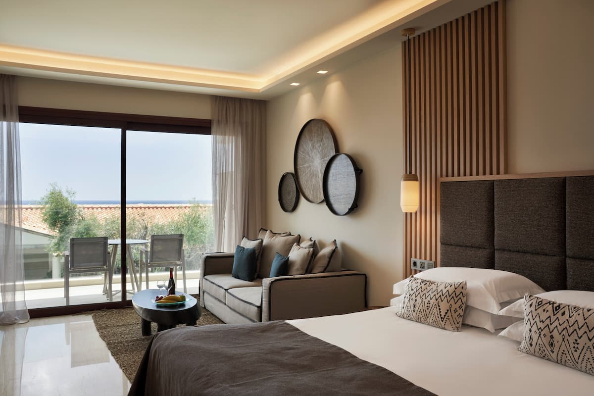 A hotel room with a bed, a sofa, a dining table, and a balcony with a view.