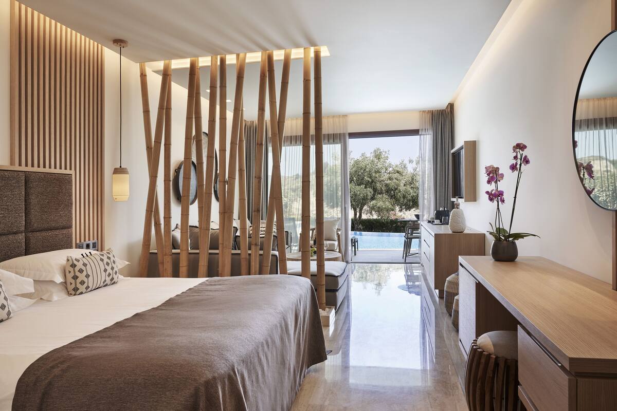 A modern hotel room with a large bed, a desk, a mirror, and a view of the outdoors.