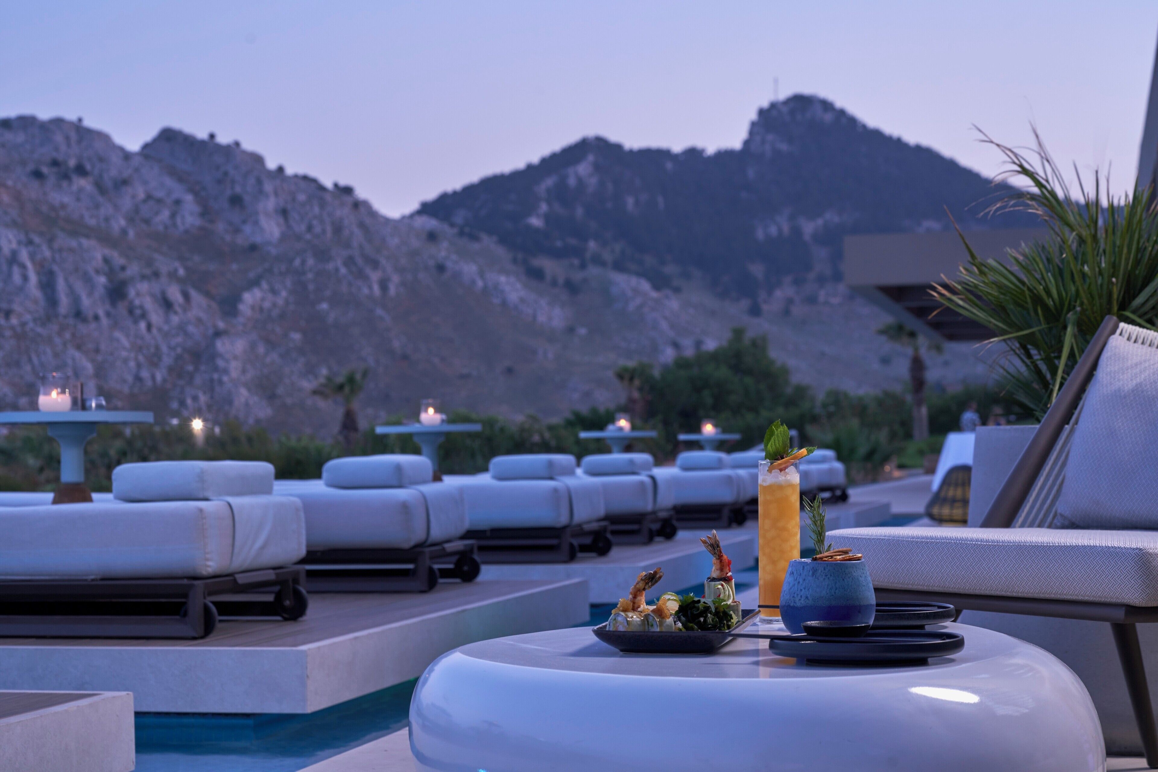 A poolside lounge area with sun loungers, a table with a drink and appetizers, and a mountainous backdrop.