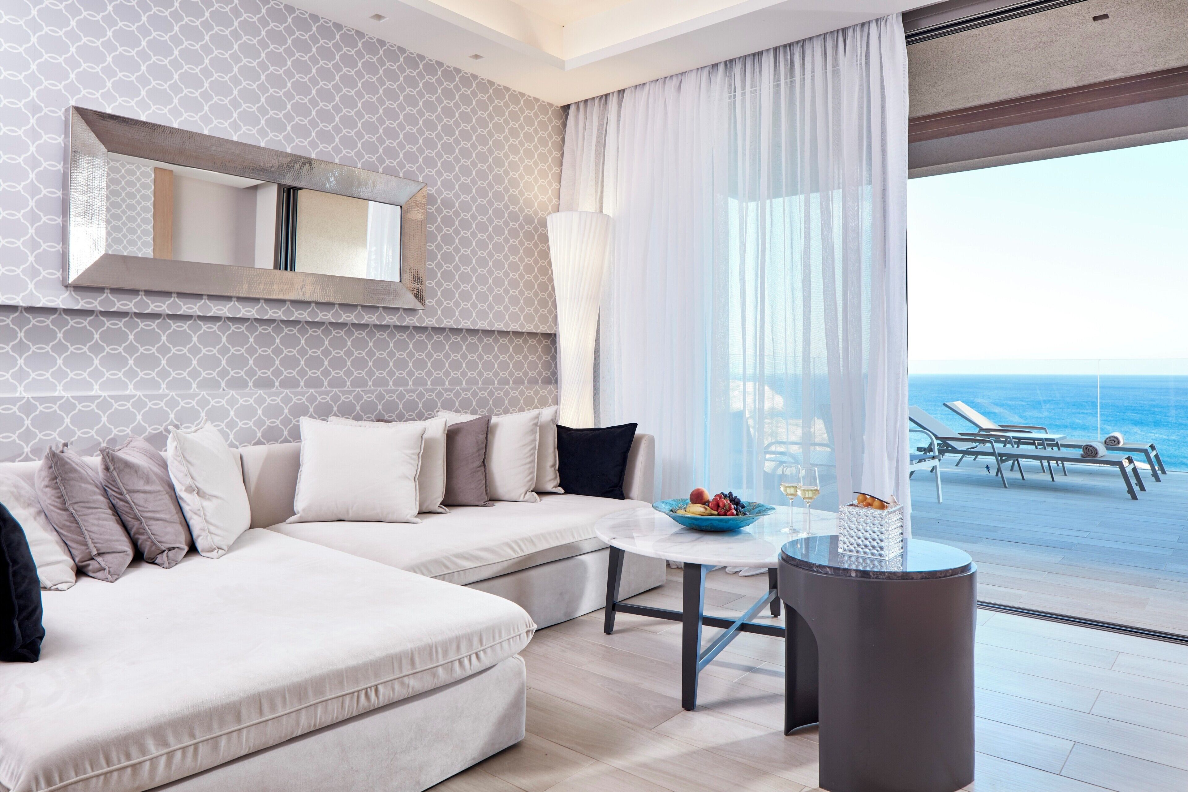 Ein modernes Hotelzimmer mit großem Fenster, einer Sitzecke, einem runden Tisch mit Obstschale und einem dekorativen Spiegel.