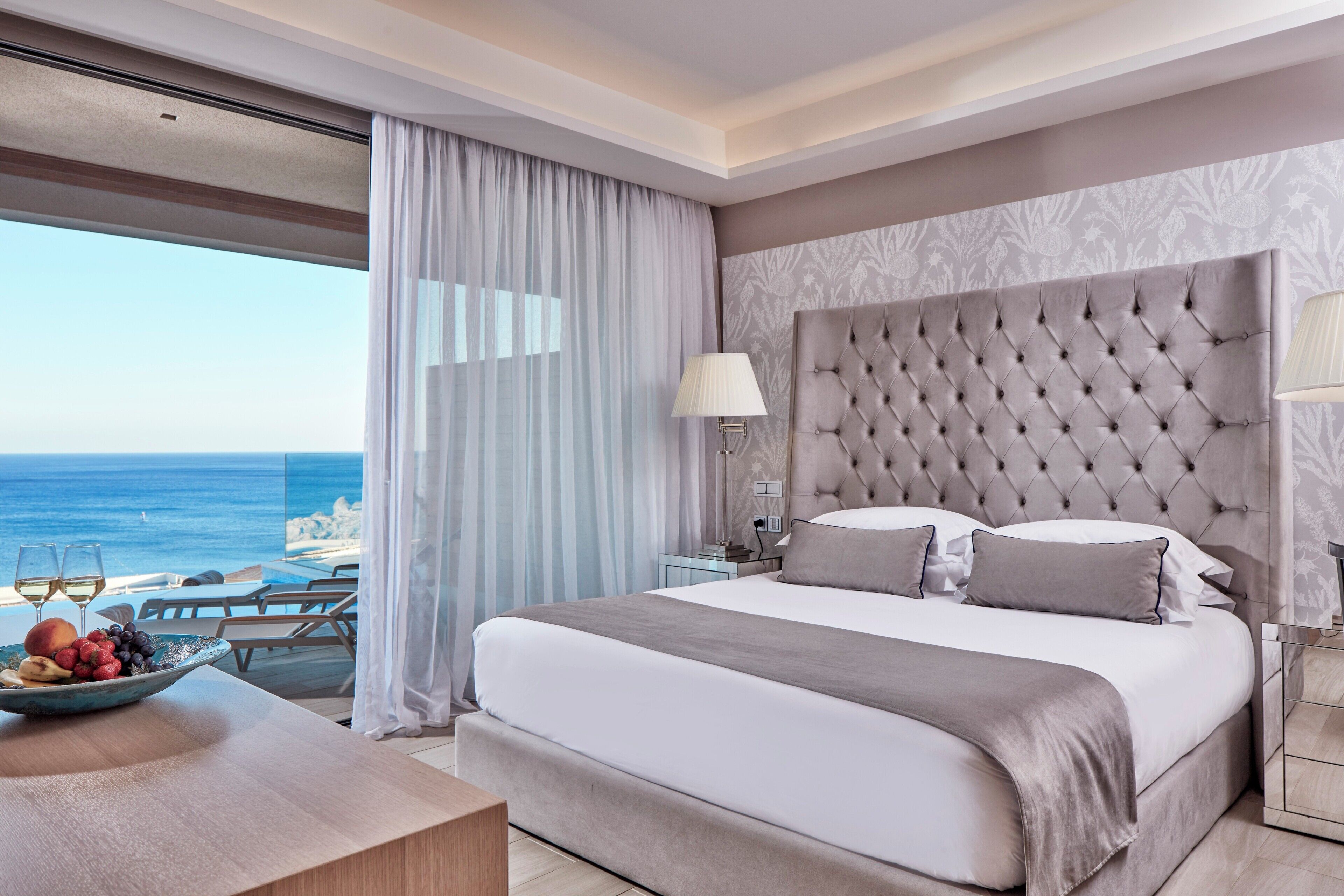 A bedroom with a large bed, a nightstand, a lamp, and a view of the ocean.