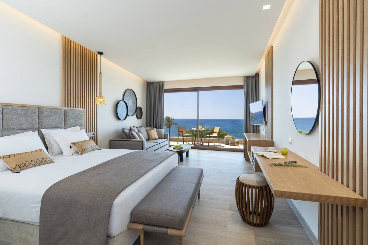 A modern hotel room with a large bed, a sofa, a dining table, and a view of the sea.