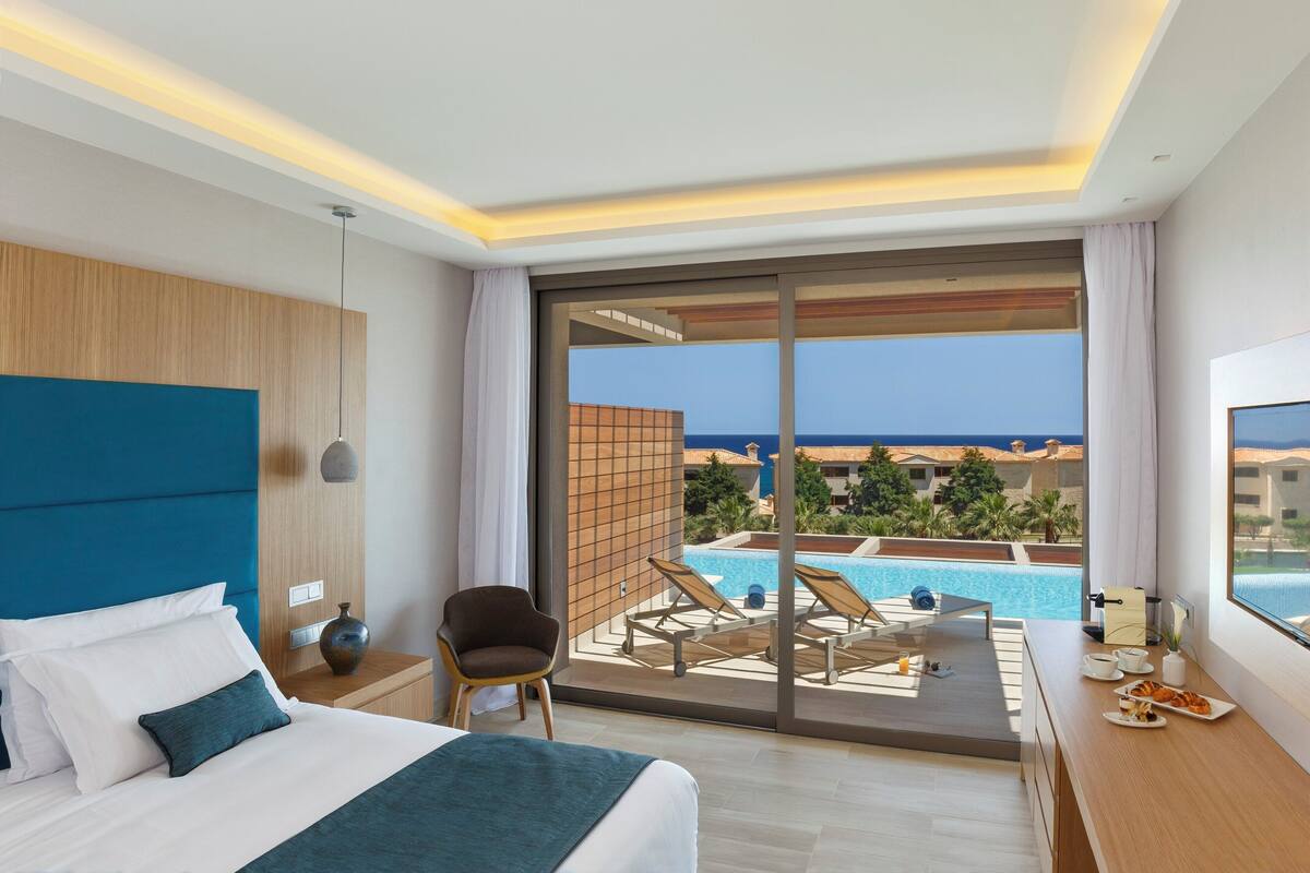 A hotel room with a bed, a chair, a dining table with breakfast, and a view of a pool and buildings.