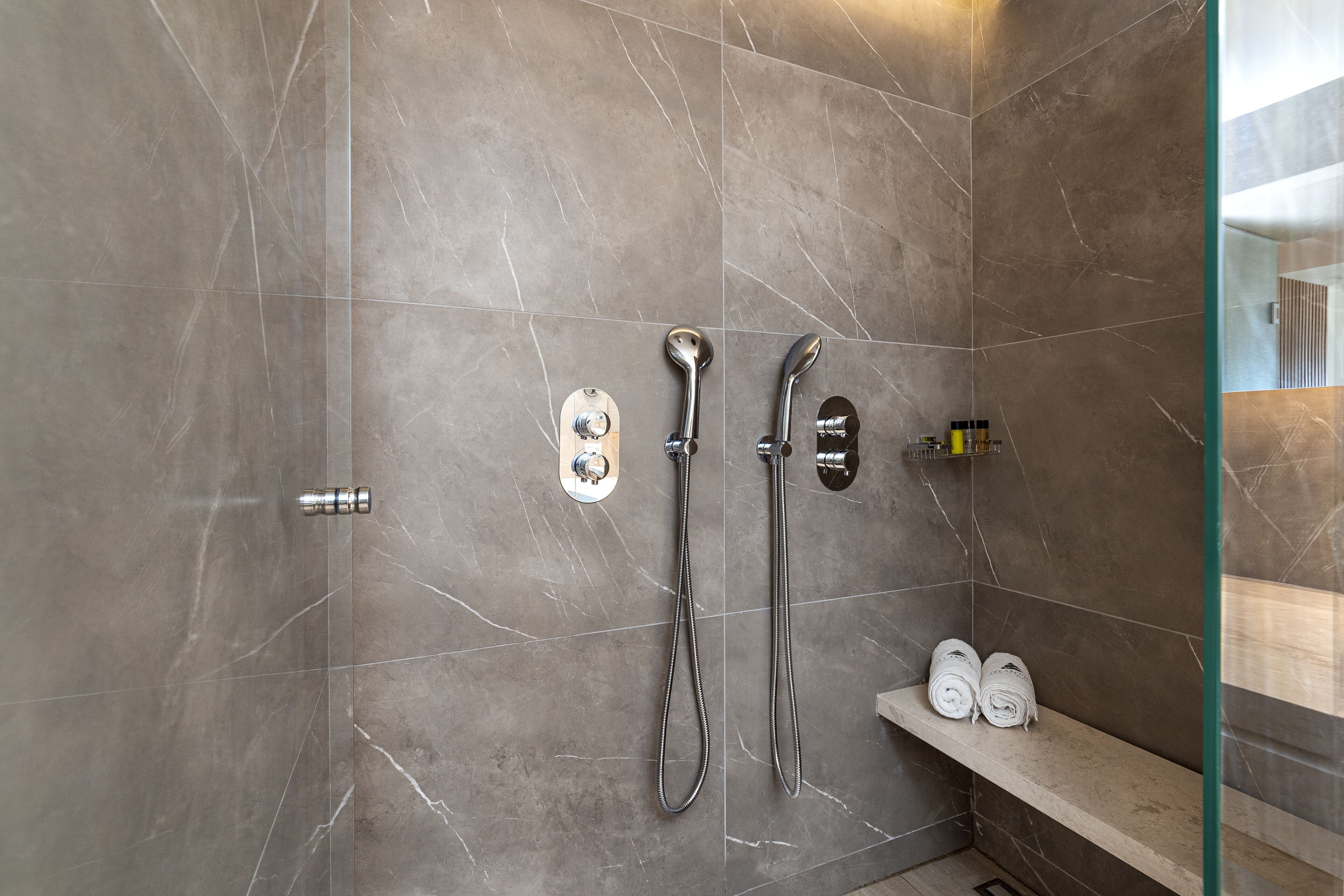 Een moderne badkamer met een douchewand van marmer, twee handdouches, een kraan op de muur en een plank met opgerolde handdoeken.